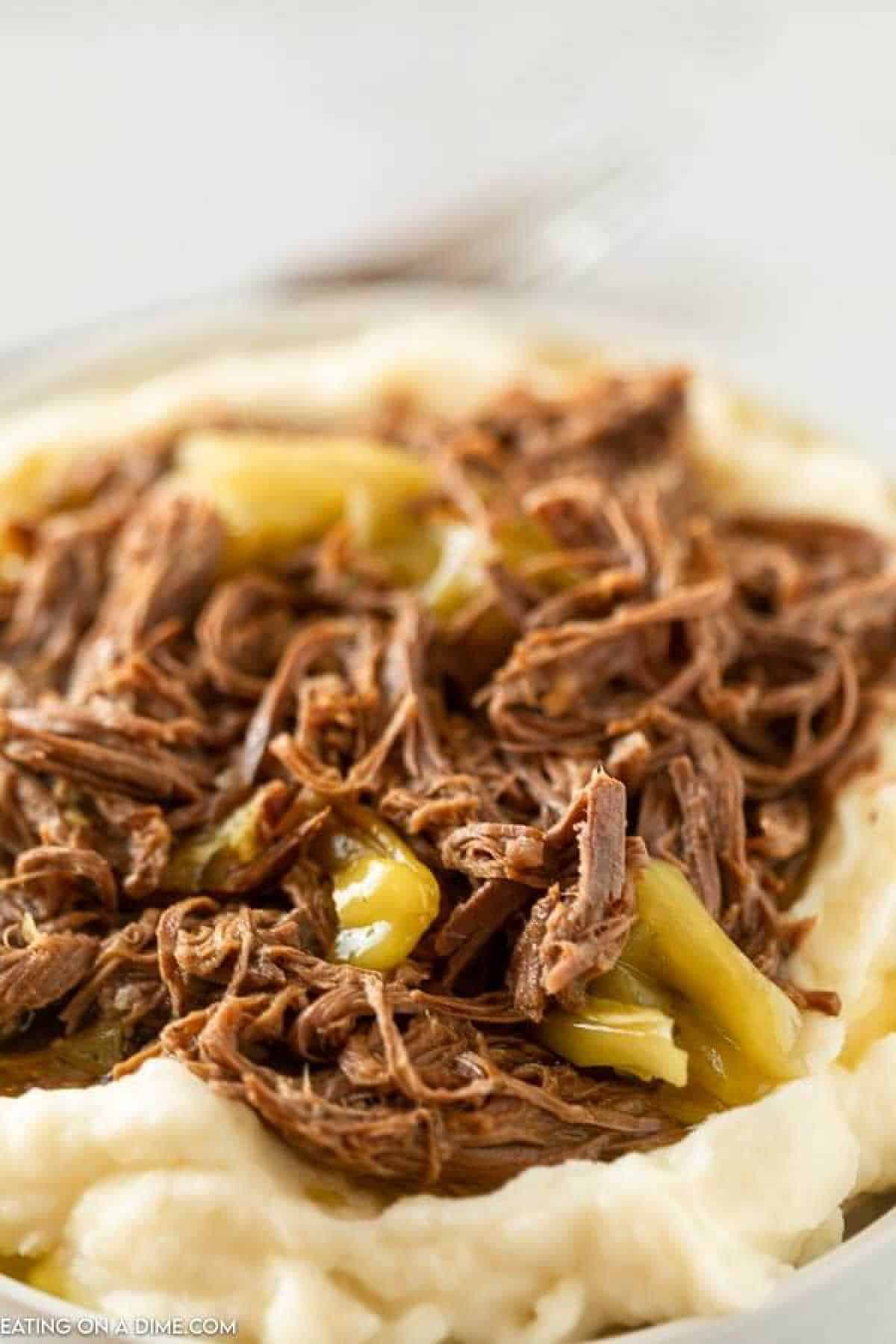 This Mississippi Pot Roast Recipe is so simple and delicious! This crock pot Mississippi Pot Roast recipes ie easy and low carb! You are going to love this original Italian Slow Cooker Mississippi Roast - it is the best and is healthy too! #eatingonadime #crockpotrecipes #slowcookerrecipes #easyrecipes A plate of creamy mashed potatoes topped with Mississippi Pot Roast, featuring tender shredded beef and sliced green peppers. The dish appears savory and appetizing, with the roast beef and peppers adding texture and color to the smooth mashed potatoes.