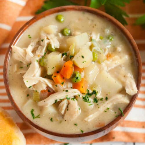 A bowl of creamy Instant Pot chicken pot pie soup with shredded chicken, potatoes, carrots, peas, and herbs, garnished with parsley. A dinner roll and striped napkin are beside the bowl.