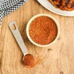 A bowl of Kickin' Chicken Seasoning sits on a wooden surface next to a metal tablespoon and a black-and-white checkered cloth. Some of the zesty spice rub is spilled from the spoon onto the wood.