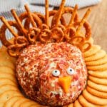 A turkey cheese ball covered in bacon bits, shaped like a turkey with pretzel sticks and mini pretzels for feathers, candy eyes, and a carrot beak, surrounded by round crackers on a wooden surface.