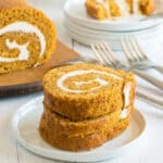 Two slices of pumpkin roll with cream cheese filling are stacked on a white plate, showcasing this delicious Pumpkin Roll Recipe, with forks and more slices on plates in the background, all set on a light wooden surface.