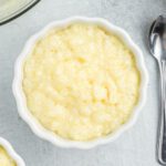 A white ramekin filled with creamy tapioca pudding sits on a light gray surface next to a metal spoon, ready to be enjoyed.