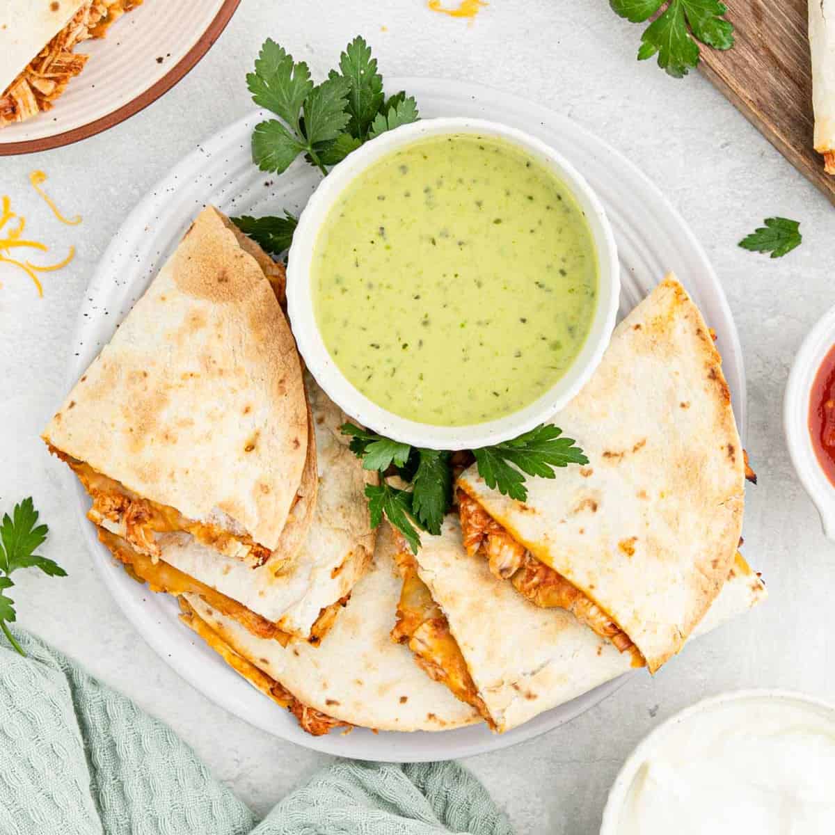 A plate of Air Fryer Chicken Quesadilla wedges is served with a bowl of creamy green sauce and garnished with fresh parsley. Other dips and a folded green napkin are nearby on a light surface.