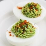 Two avocado deviled eggs on a white plate, filled with a creamy green avocado mixture, garnished with chopped green onions and a sprinkle of red paprika.