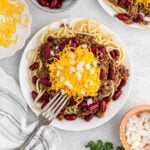 A plate of spaghetti topped with chili, kidney beans, shredded cheddar cheese, and chopped onions sits on a table with a fork—a classic Cincinnati Chili Recipe—surrounded by bowls of cheese and onions.