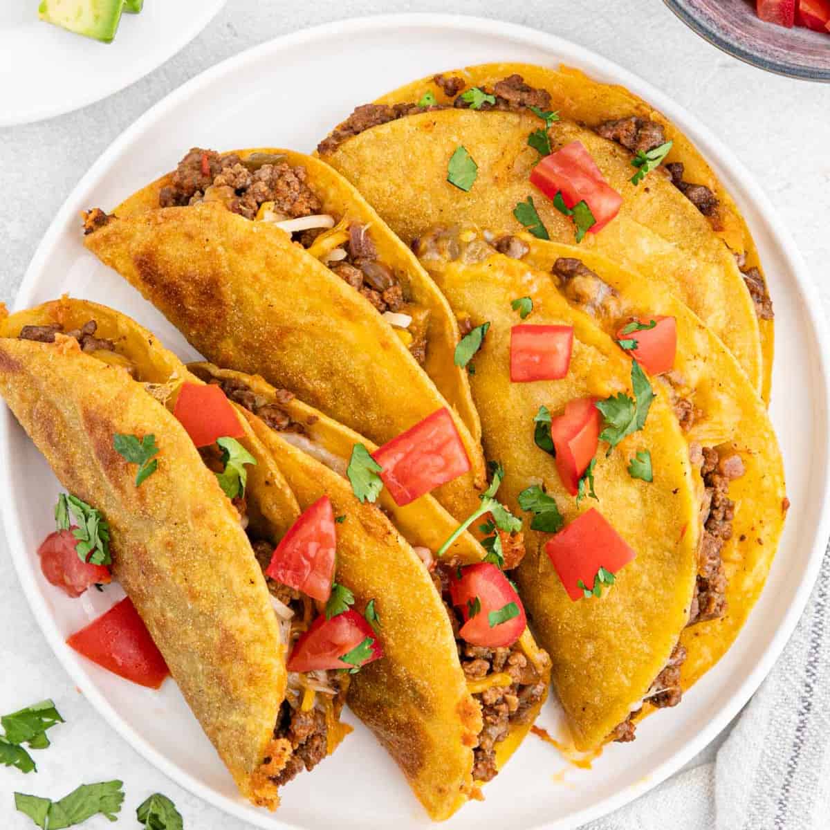 A plate of four crispy tacos made with our Fried Tacos Recipe, filled with seasoned ground beef, shredded cheese, and topped with chopped tomatoes and fresh cilantro.