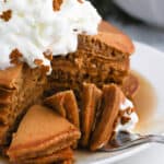 A stack of fluffy Gingerbread Pancakes topped with whipped cream and small gingerbread-shaped sprinkles, with a slice cut out and served on a fork, sits on a plate drizzled with syrup.