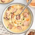 A bowl of creamy Kielbasa Potato Soup with sliced sausage, diced potatoes, carrots, and herbs sits invitingly, a spoon inside. Nearby are a small dish of red pepper flakes and part of a napkin.