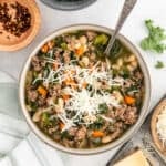 A bowl of hearty Sausage Kale White Bean Soup with carrots and celery, topped with shredded cheese. A spoon rests in the bowl, while grated cheese and red pepper flakes are scattered nearby on the table.