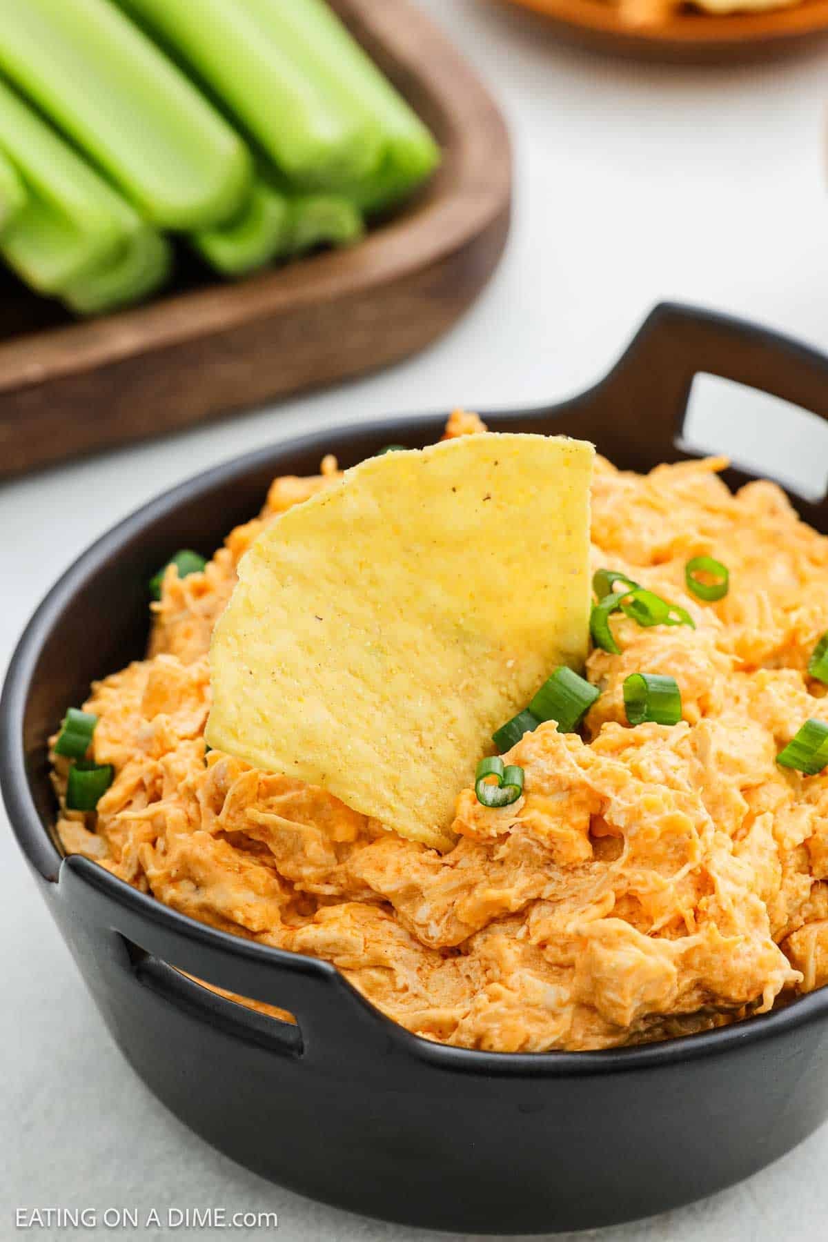 A black bowl filled with creamy Crock Pot Buffalo Chicken Dip, topped with chopped green onions and a tortilla chip, with celery sticks visible in the background.