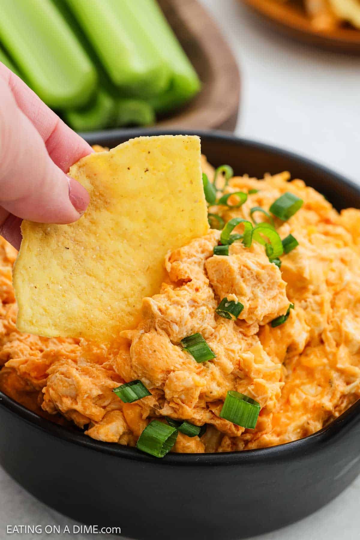 A hand dips a tortilla chip into a bowl of creamy Crock Pot Buffalo Chicken Dip topped with chopped green onions. Celery sticks are visible in the background.