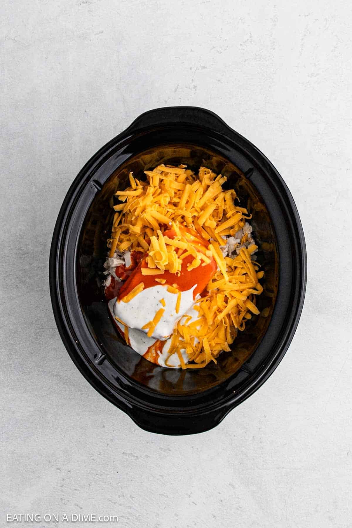 A black slow cooker filled with shredded cheddar cheese, creamy white sauce, and zesty orange sauce atop other ingredients, perfect for making Crock Pot Buffalo Chicken Dip, sits on a light-colored countertop.