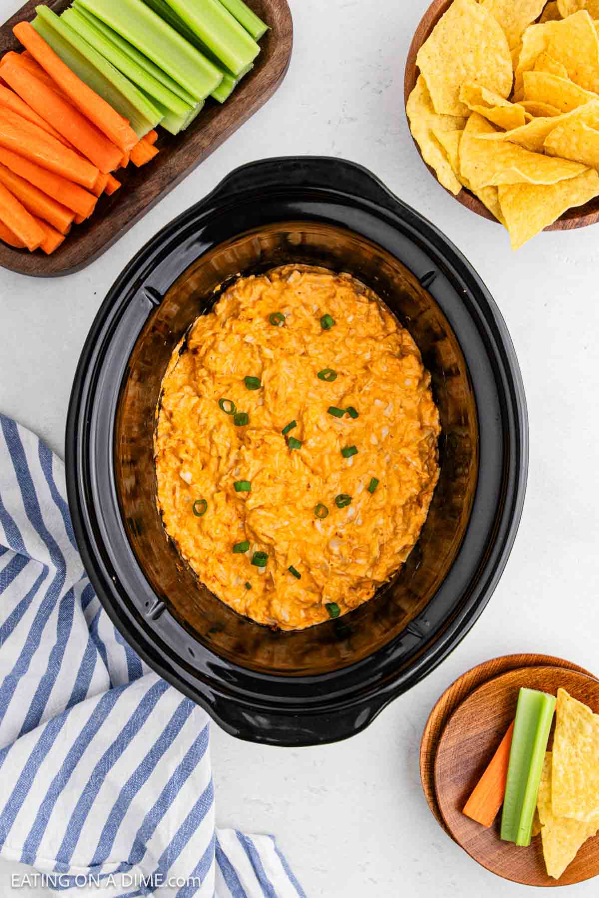 A Crock Pot Buffalo Chicken Dip sits creamy and topped with sliced green onions, surrounded by bowls of tortilla chips, celery, and carrots on a white surface. A blue and white striped cloth is nearby.