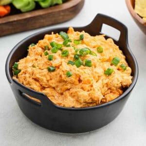 A black bowl filled with creamy Crock Pot Buffalo Chicken Dip, topped with chopped green onions, sits on a light surface with vegetables and chips nearby.