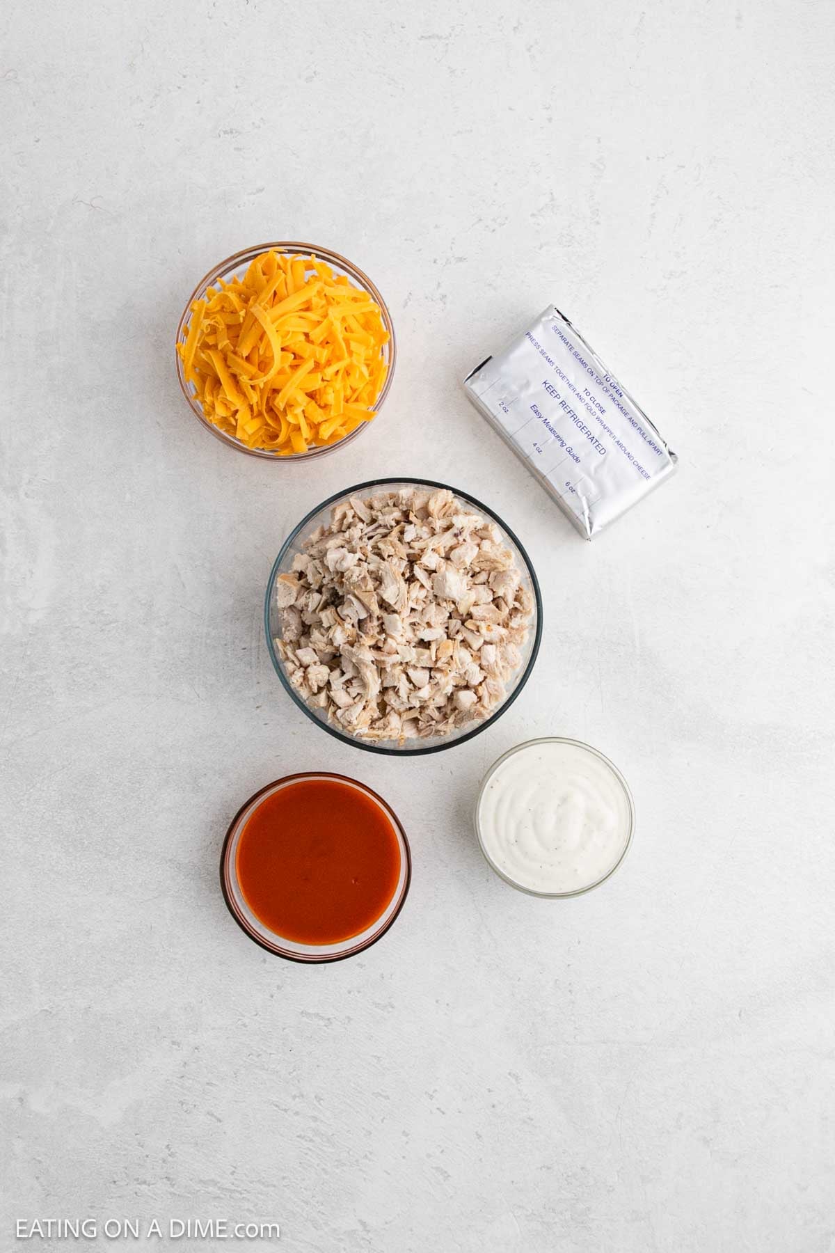 Five ingredients for Crock Pot Buffalo Chicken Dip on a white surface: a bowl of shredded cheddar cheese, a block of cream cheese, chopped cooked chicken, buffalo sauce, and ranch dressing.