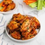 A plate of crock pot buffalo chicken wings glazed to perfection and garnished with herbs, with celery and carrot sticks in the background on a white surface. A second plate with wings, carrots, and celery is also visible.