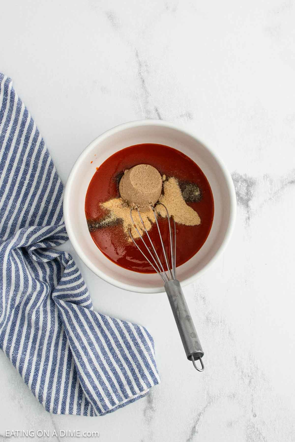 A bowl filled with ketchup, brown sugar, and spices—perfect for crock pot buffalo chicken wings—is being mixed with a whisk. A blue and white striped kitchen towel lies next to the bowl on a white marble surface.