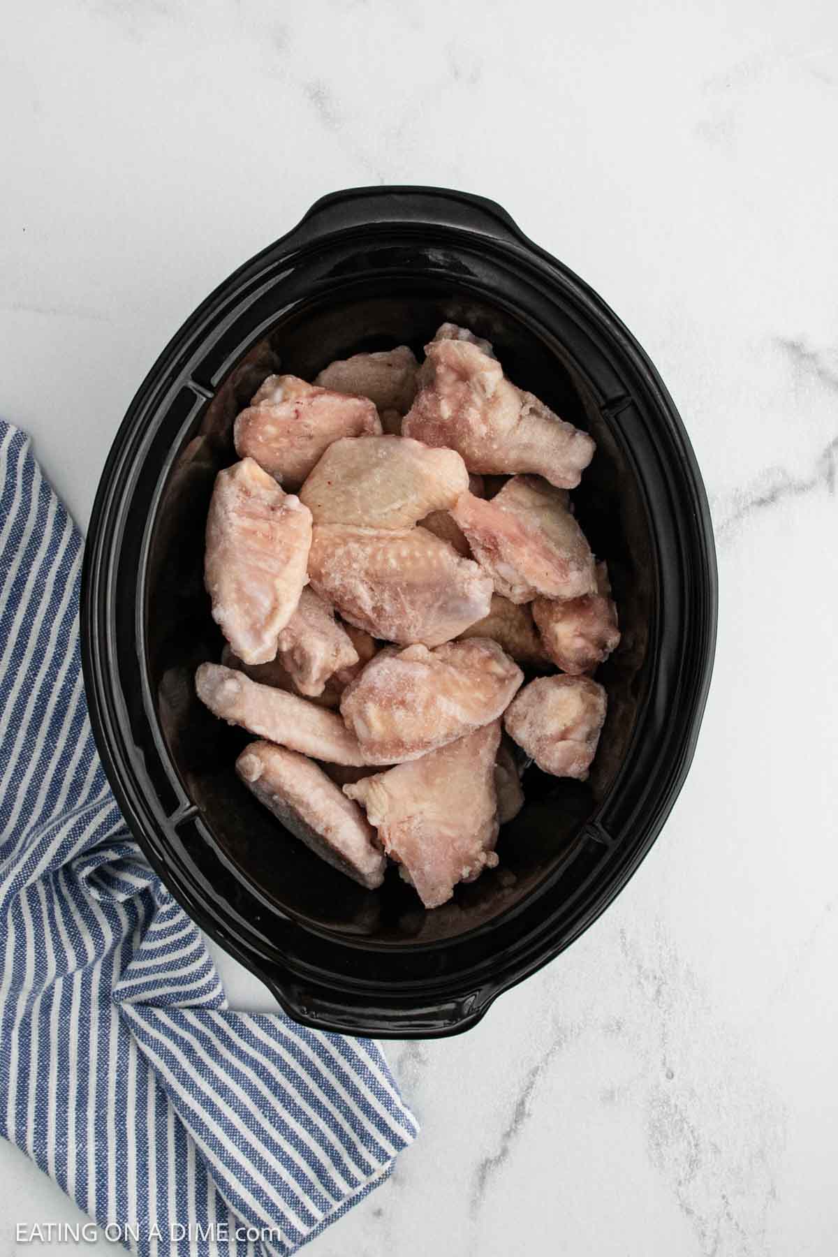 Raw crock pot buffalo chicken wings placed inside a black slow cooker on a white marble surface, with a blue and white striped kitchen towel next to it.