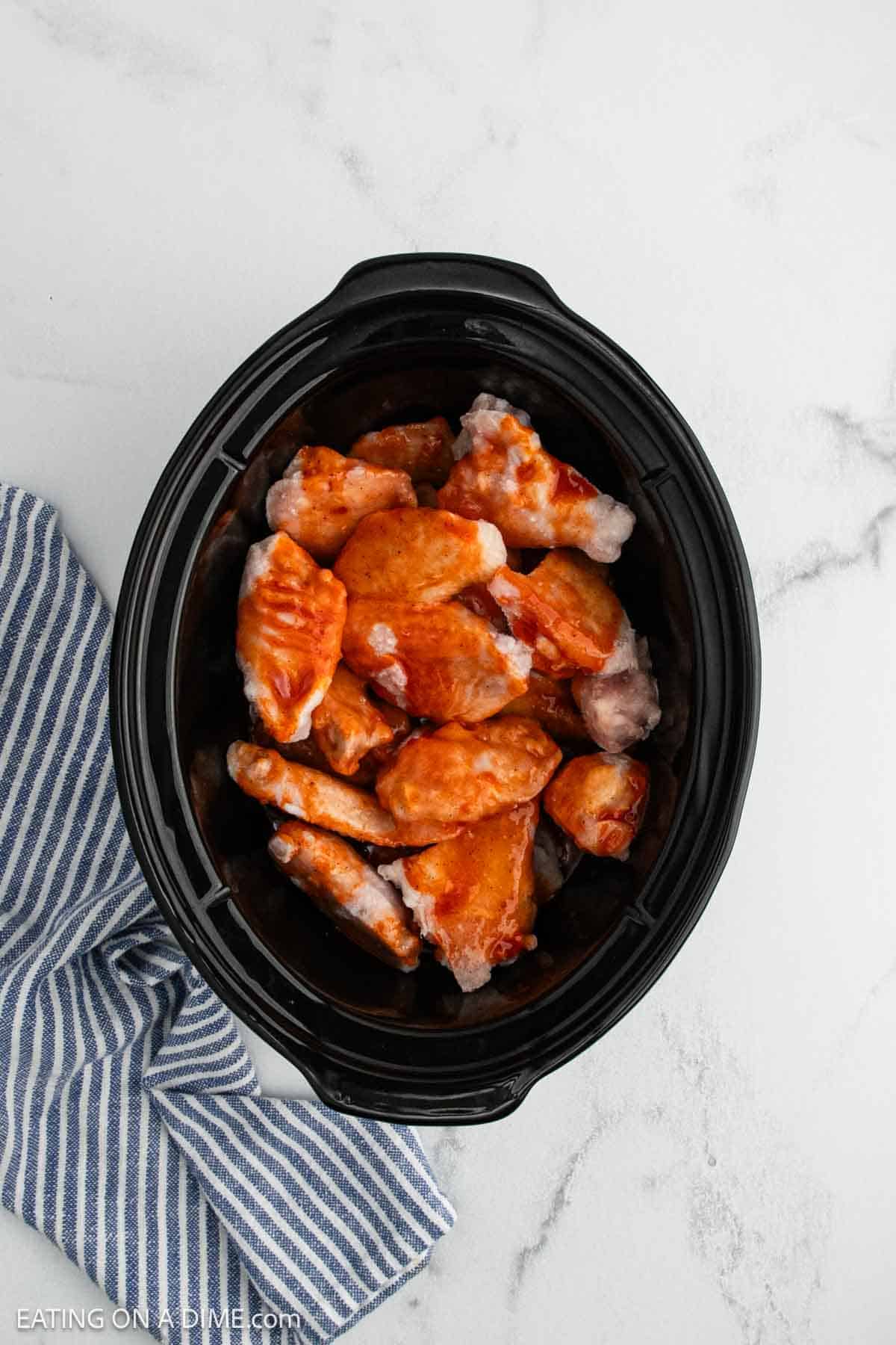 A black crock pot filled with raw buffalo chicken wings coated in sauce sits on a white marble surface, next to a blue and white striped cloth.