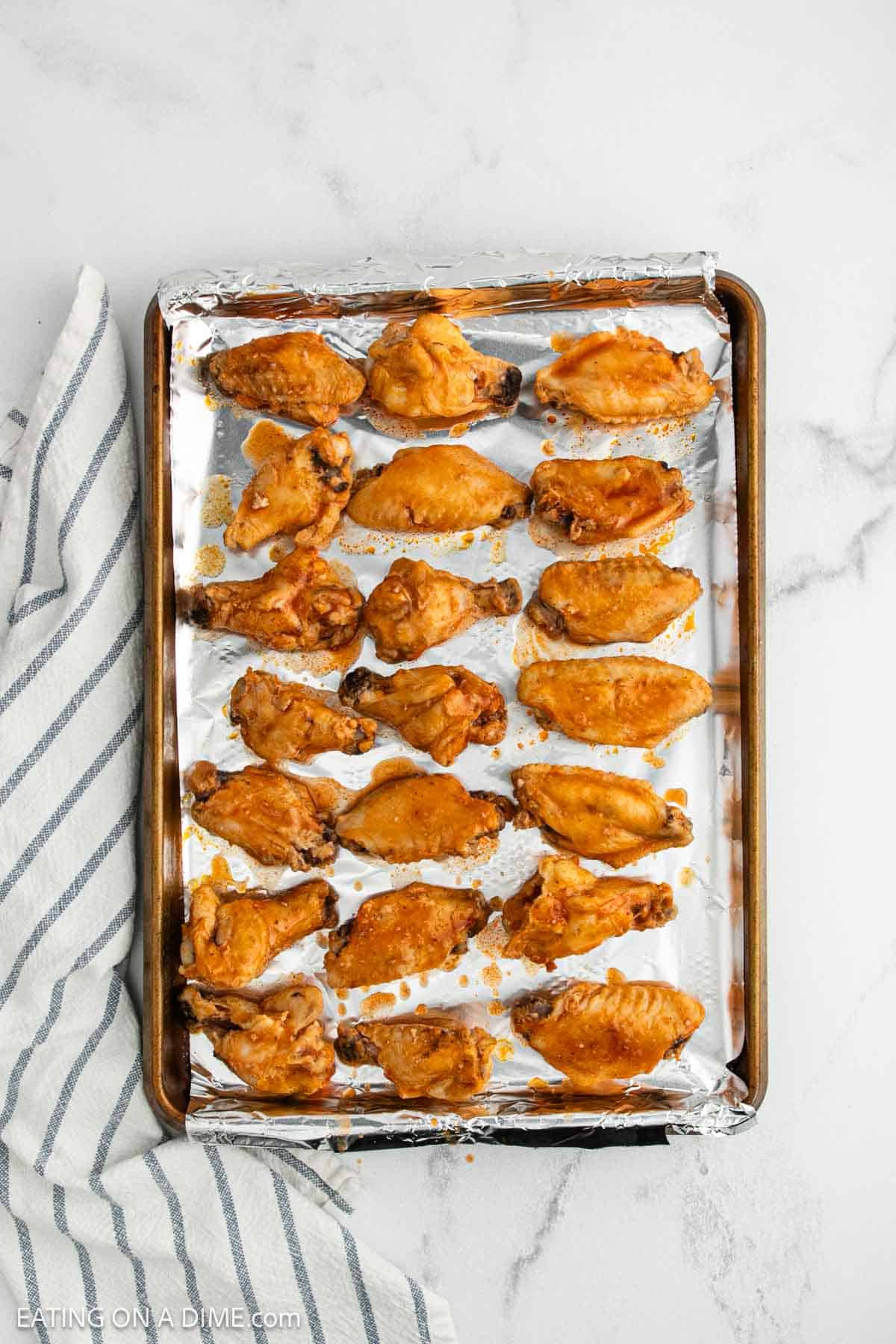 A baking sheet lined with foil holds rows of crock pot buffalo chicken wings coated in sauce. The sheet rests on a white marble surface next to a blue-and-white striped towel.