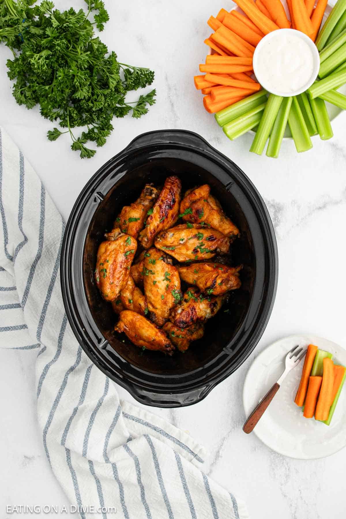 A crock pot of buffalo chicken wings sits on a marble surface. Nearby are fresh parsley, a plate of celery and carrot sticks with ranch dip, and a striped kitchen towel.