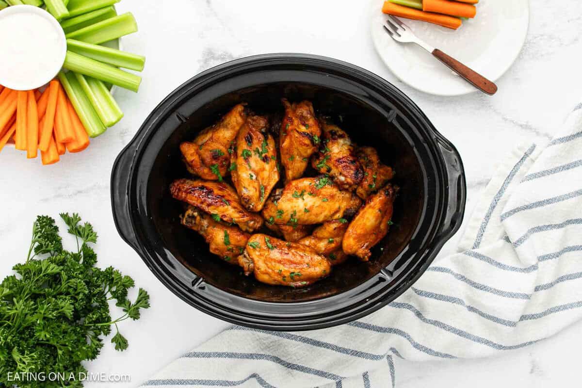 A crock pot filled with buffalo chicken wings garnished with herbs sits on a white surface. Fresh celery and carrot sticks, parsley, a fork, and a striped towel are arranged nearby for the perfect game day spread.
