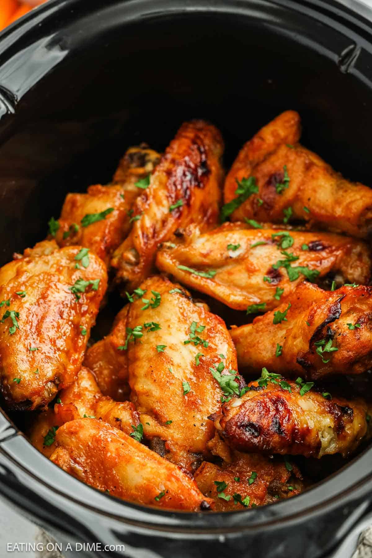 Crock pot buffalo chicken wings coated in sauce and garnished with chopped parsley are piled inside a black slow cooker. The wings appear to be cooked, with a golden-brown and slightly crispy texture.
