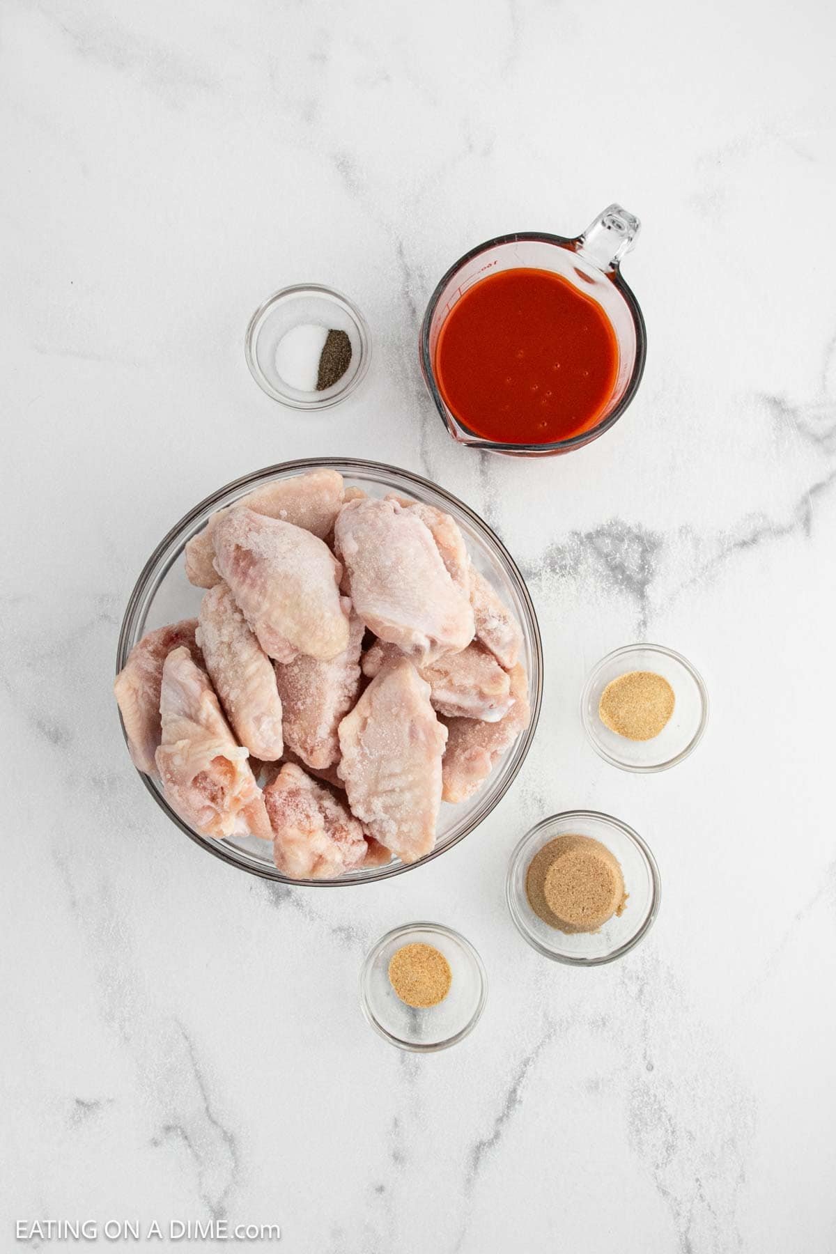 A glass bowl of raw chicken wings, perfect for crock pot buffalo chicken wings, surrounded by small bowls of spices, a glass cup of red sauce, and a pinch of black pepper on a white marble surface.