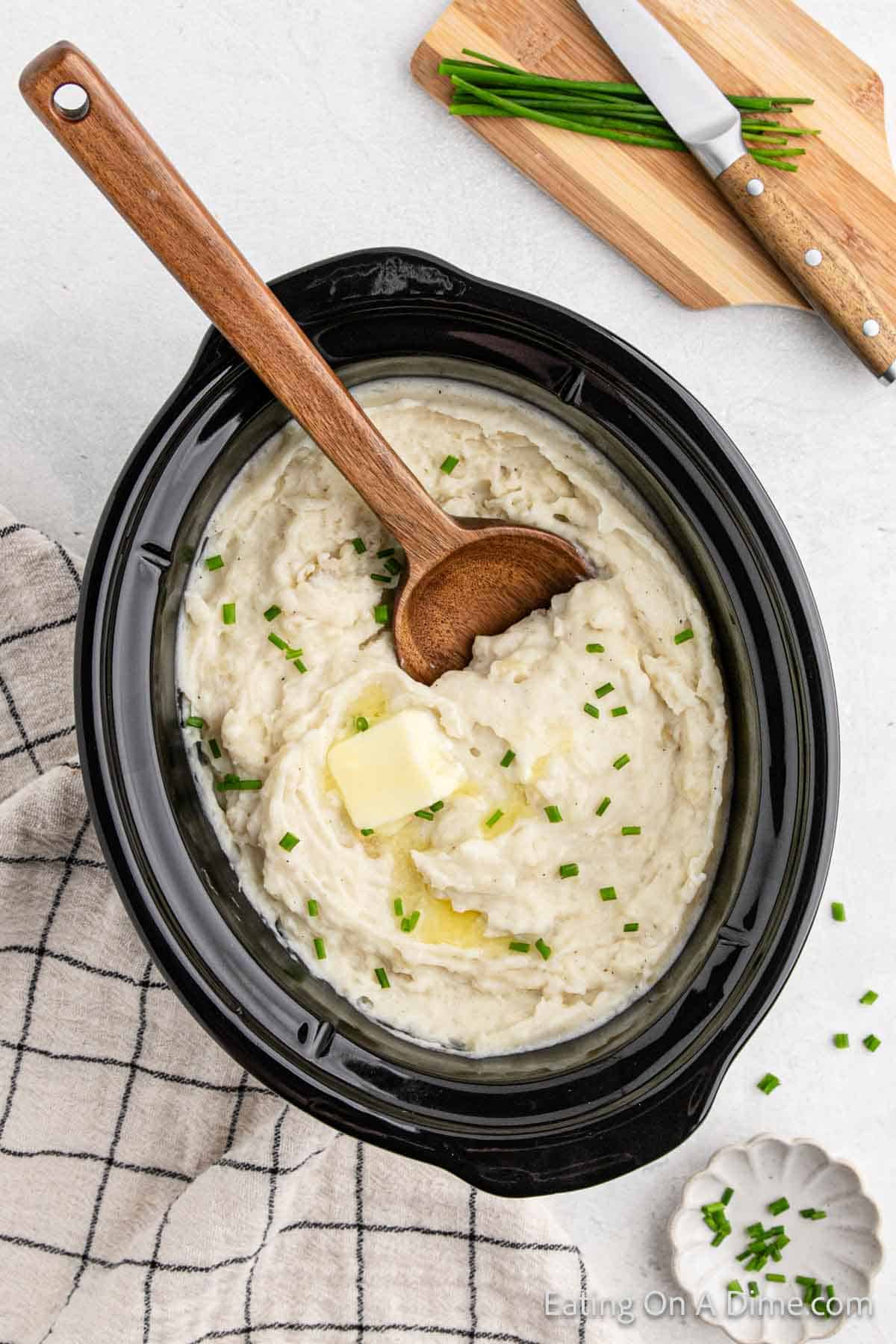Mashed Potatoes in the crock pot with a wooden spoon and topped with melted butter and chives