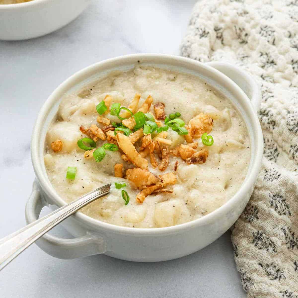 A bowl of creamy Crockpot Potato Soup garnished with chopped green onions and crispy fried onions, with a spoon in the bowl. A patterned cloth napkin is beside the bowl.
