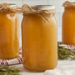 A glass mason jar filled with light brown Chicken Broth Substitutes, sealed with a metal lid and decorated with a twine bow. Fresh rosemary sprigs and striped cloths are arranged nearby on a white surface.