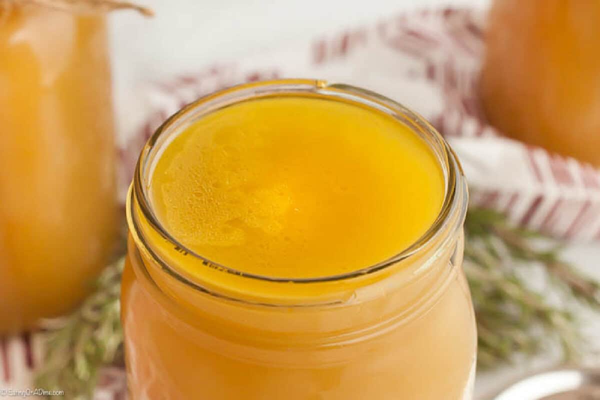 A close-up of an open glass jar filled with golden-yellow ghee, showing its smooth, glossy surface. In the background, blurred jars and a striped cloth hint at kitchen staples like Chicken Broth Substitutes.