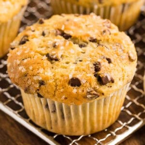 A freshly baked chocolate chip muffin, part of a batch of irresistible Chocolate Chip Muffins, is topped with coarse sugar and rests on a wire cooling rack, with more muffins tempting in the background.