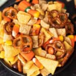 A bowl filled with a halloween chex mix featuring pretzels, cereal squares, candy corn, and chocolate pieces.