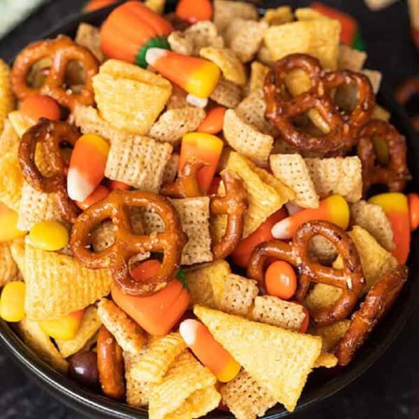 A bowl filled with a halloween chex mix featuring pretzels, cereal squares, candy corn, and chocolate pieces.