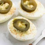 Three Jalapeño Deviled Eggs, topped with sliced pickled jalapeños and a sprinkle of paprika, are arranged on a white plate, with a small metal fork visible to the side.