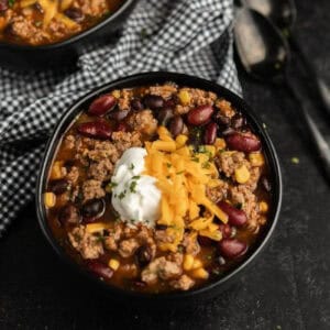 A black bowl filled with chili inspired by a crockpot taco soup recipe—ground beef, kidney and black beans, corn, cheddar cheese, sour cream, and herbs—served with a checkered cloth and spoons nearby.