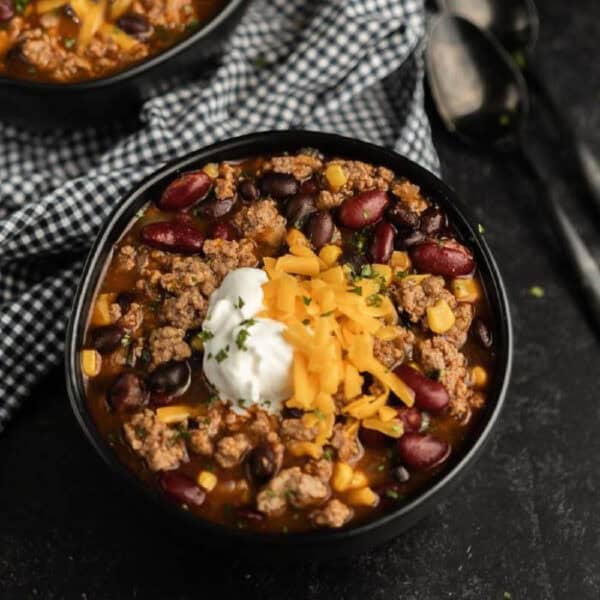 A black bowl filled with chili inspired by a crockpot taco soup recipe—ground beef, kidney and black beans, corn, cheddar cheese, sour cream, and herbs—served with a checkered cloth and spoons nearby.