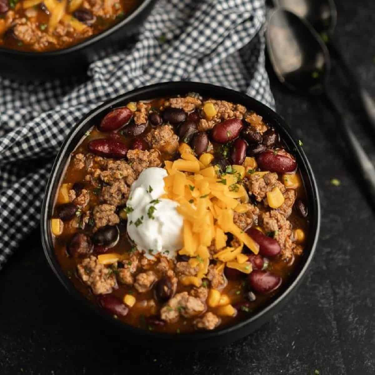 A black bowl filled with chili inspired by a crockpot taco soup recipe—ground beef, kidney and black beans, corn, cheddar cheese, sour cream, and herbs—served with a checkered cloth and spoons nearby.