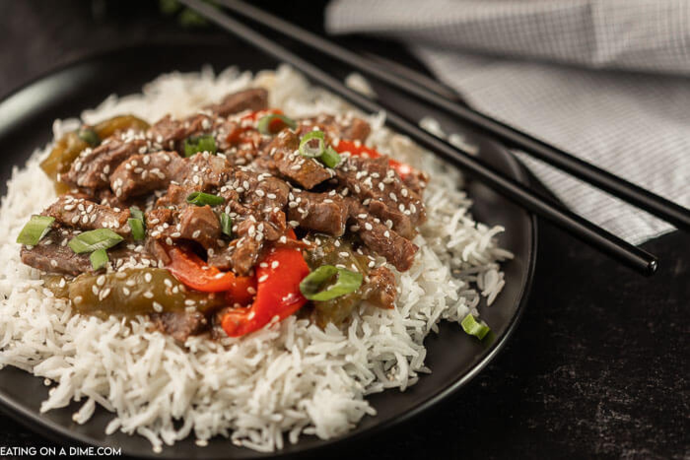 A plate of this pepper steak over rice on a black plate with chop sticks next to the plate. 