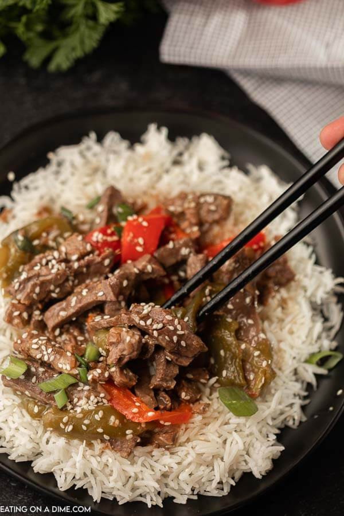 Need an easy crock pot recipe? This Crockpot Pepper Steak Recipe is delicious! Easy slow cooker pepper steak and rice recipe is simple to make and delicious. This Chinese pepper steak recipe the best healthy beef stir fry recipe and is one of my favorite slow cooker easy recipes. You will love this Eating on a Dime Recipe! #eatingonadime #crockpotrecipes #steakrecipes #beefrecipes A plate filled with rice and then this crock pot pepper steak on top with chop sticks picking up some of the pepper steak.