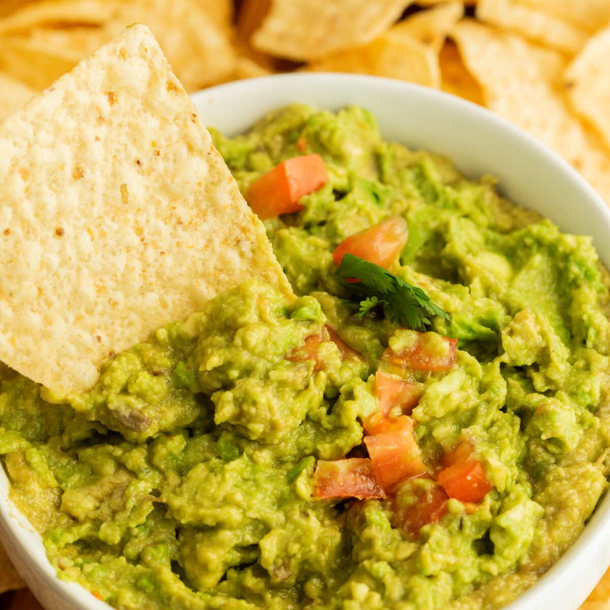 Guacamole in a bowl topped with diced tomatoes with a chip dipped into the guacamole and tortilla chips on the side