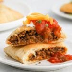 Two halves of taco pockets filled with seasoned ground beef, topped with salsa, sour cream, shredded cheddar cheese, and chopped herbs, served on a white plate.