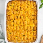 A Chicken Tater Tot Casserole in a white dish, topped with crispy tater tots, melted cheddar cheese, and garnished with chopped green herbs.