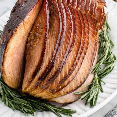 Sliced slow cooker Christmas ham arranged on a white plate, garnished with fresh rosemary sprigs. The ham has a shiny, caramelized crust and juicy, tender slices.