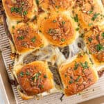 Golden, cheesy French Dip Sliders pull apart easily, topped with chopped herbs and crispy onions on a baking tray. The melted cheese stretches between the soft, shiny rolls.