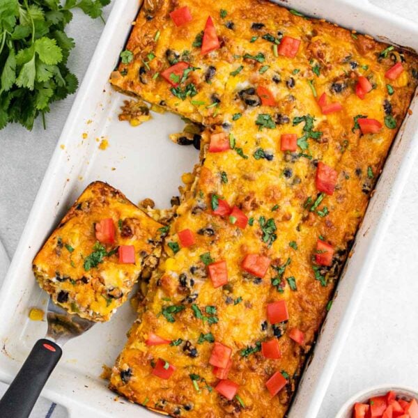 A Mexican breakfast casserole topped with melted cheese, chopped tomatoes, and herbs sits in a white dish, with one square piece being served on a spatula. Fresh parsley and a bowl of diced tomatoes are nearby.
