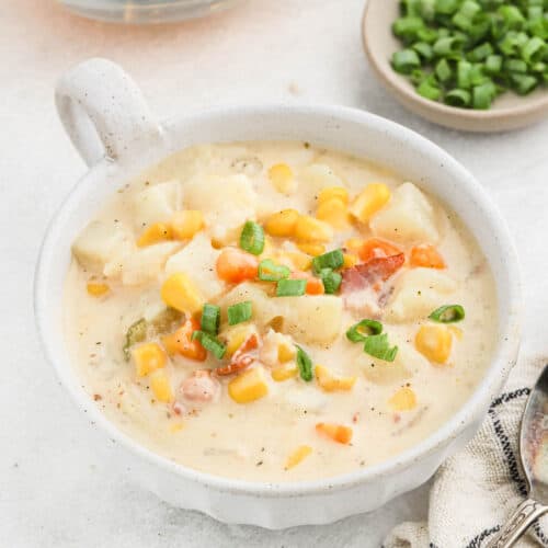 A creamy potato corn chowder in a white bowl, filled with corn, potatoes, and carrots, and garnished with chopped green onions. A small plate of extra green onions is in the background.
