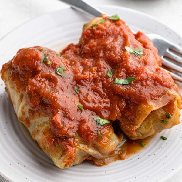 Two slow cooker cabbage rolls stuffed with filling, topped with chunky tomato sauce and garnished with small bits of green herbs, served on a white plate with a fork beside them.