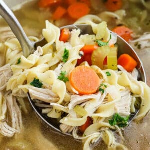 A ladle lifting homemade chicken noodle soup with spiral noodles, shredded chicken, sliced carrots, celery, and parsley from a pot of flavorful broth.